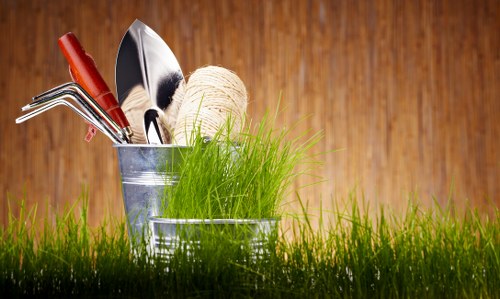 A close-up view of two small, round metal garden scoops filled with gardening tools and supplies, including a trowel, cultivator, and a ball of twine, all set against a backdrop of fresh green grass growing outdoors. The tools are arranged with some handles visible, displaying natural wood and red plastic grips. The surface beneath the grass appears to be part of a well-maintained garden or outdoor space, possibly at a residential property in Southgate. The scene is illuminated by natural daylight, highlighting the vibrant green of the grass and the metallic shine of the gardening tools, suggesting a pleasant, clear day suitable for gardening activities. The image emphasizes gardening preparations or maintenance tasks typical of services provided by Gardening Services Southgate, involving planting, cultivation, or general garden care, with the focus on outdoor garden elements and tools used for such work.