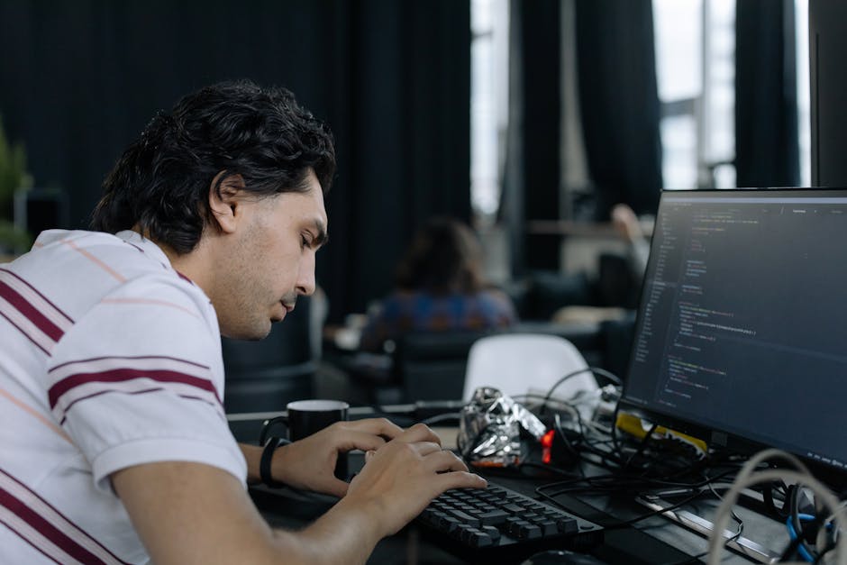 A silver laptop placed on a wooden table displaying a screen filled with lines of code, with a closed orange pen resting beside it. The background is softly blurred, showing a cozy indoor space with w